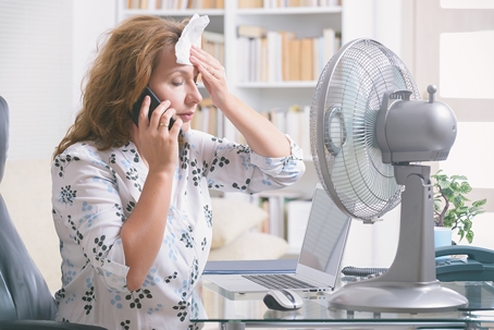 woman in front of fan, on the phone as she wipes sweat of her forehead.